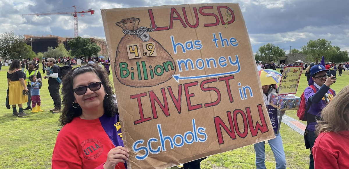 A woman in a red UTLA t-shirt holds a cardboard sign with red, blue, white and green lettering showing a big bag of money and saying “LAUSD has the money, 4.9 billion, invest in schools now.” She’s standing in a field with other demonstrators in the background. 