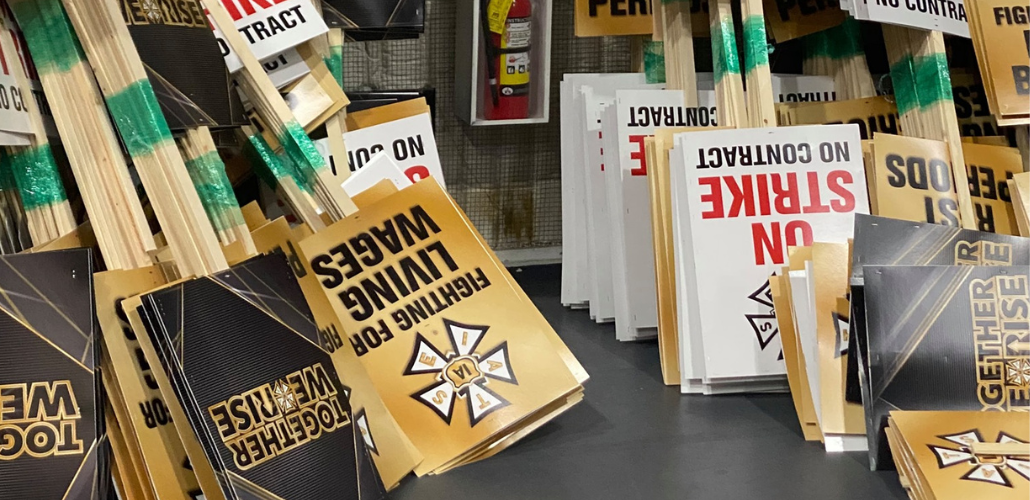 Picket signs stacked against a wall. They say "FIGHTING FOR LIVING WAGES," "ON STRIKE NO CONTRACT," and "TOGETHER WE RISE" and have the IATSE logo. There is also a fire extinguisher in the photo.