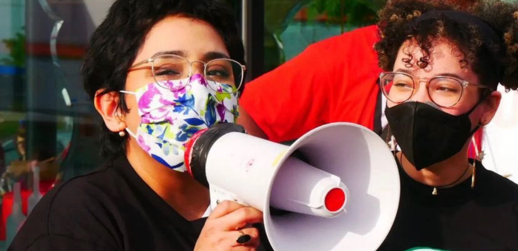 One masked worker speaks into a bullhorn as another looks on. Both look happy and young.