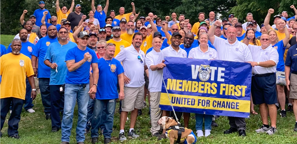 Big crowd stands on grass outdoors on a sunny day behind a banner that reads "Vote Members First, United for Change." Everyone is wearing a union shirt: white, blue, or yellow. Many have fists in the air and everyone is smiling. Someone in front has a dog on a leash.