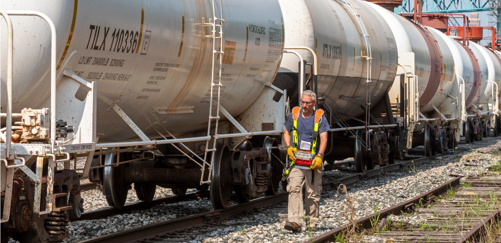 A worker in reflective vest walks alongside a train, holding a remote control device