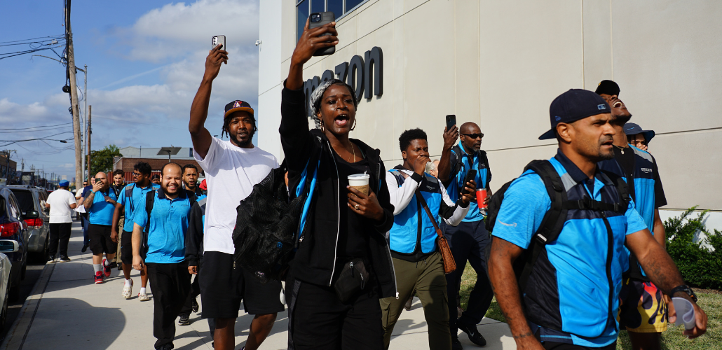 Workers, some in blue vests, march outside along an Amazon building. Some are holding their phones aloft, recording. Many are Black. They look like they are striding confidently and chanting. 