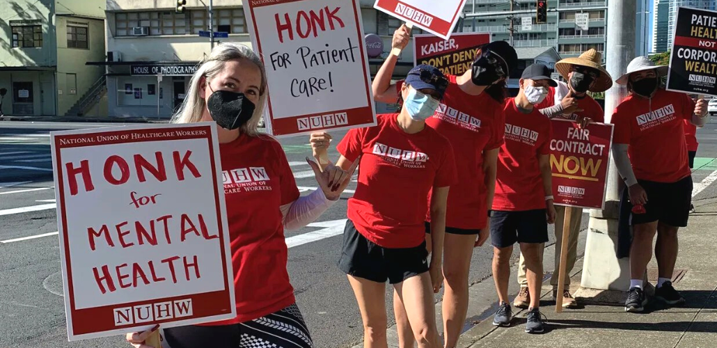 Six picketers in red NUHW T-shirts stand on outdoors along a sidewalk. Most are masked. Some carry handmade signs that say "Honk for mental health" and "honk for patient care." Others carry printed signs that say "Fair contract now" and "Patient health not corporate wealth."