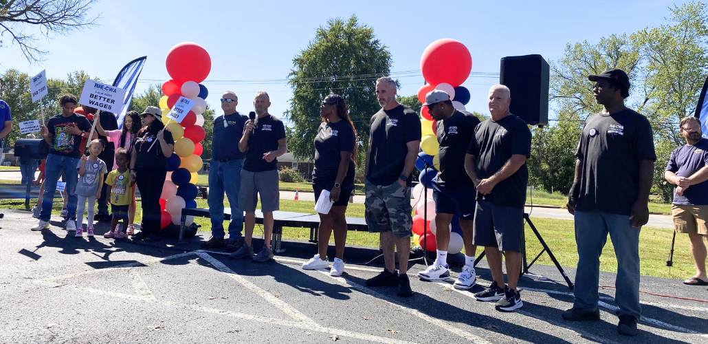 About a dozen adults and a couple children, white and Black, men and women, stand mostly silhouetted in bright sun. Most wear black union T-shirts.They're outdoors, on pavement in front of grass. One holds a mic. Some hold printed signs that say, "IUE-CWA, Better Wages." There are also two columns of balloons, each a spiral design in white, red, yellow, and black.