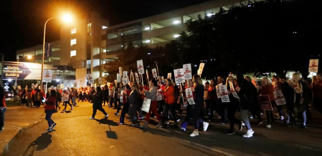 Workers with "On Strike" picket signs march past a hospital in the near-dark, silhouetted by one bright light.