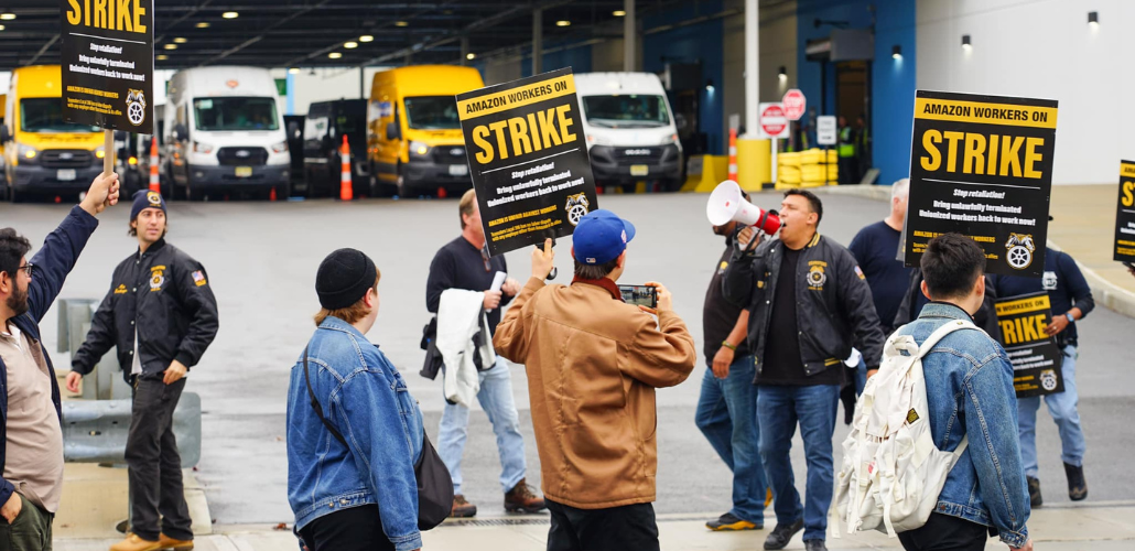 Workers picket in front of an Amazon facility where delivery vehicles are visible. Some carry printed "Amazon workers on strike" signs with a Teamsters logo. Others wear satin jackets with a Teamsters logo, and one shouts into a bullhorn.