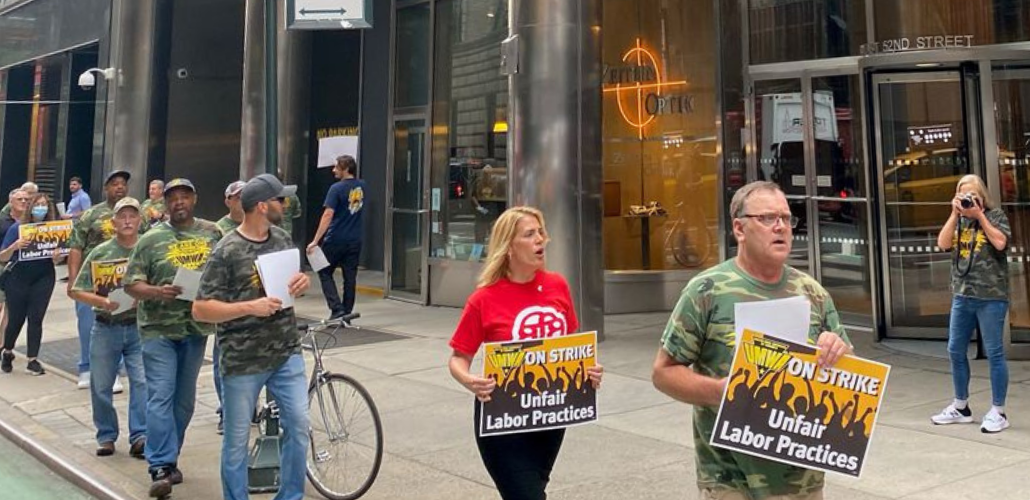 Picketers march outside an office building, most in camouflage shirts. They carry signs: "UMWA ON STRIKE: Unfair Labor Practices"