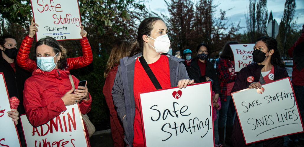 Three women dressed in red hold hand-lettered signs: "Safe Staffing Saves Lives" and "Patients Over Profits"