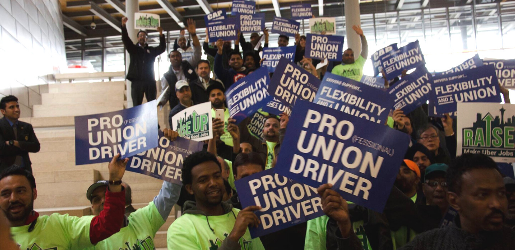 A big crowd of workers in matching green T-shirts hold up printed signs reading "PRO(FESSIONAL) UNION DRIVER"