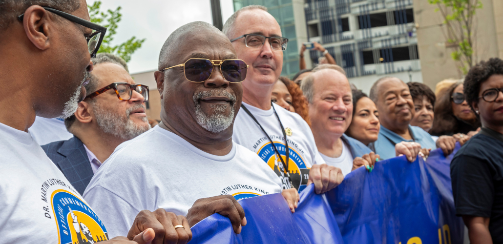 Black and white people, including UAW President Shawn Fain, hold a blue banner (text not visible). They are wearing matching T-shirts for a Martin Luther King Official Commemoration.