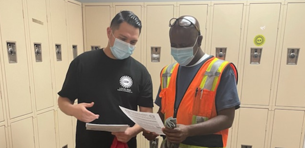 Two men talk inside an auto plant locker room, looking together at leaflets. Both wear masks and one wears an orange reflective vest.