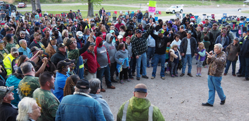 A crowd of people forms a big circle outdoors. One person stands in the middle speaking.