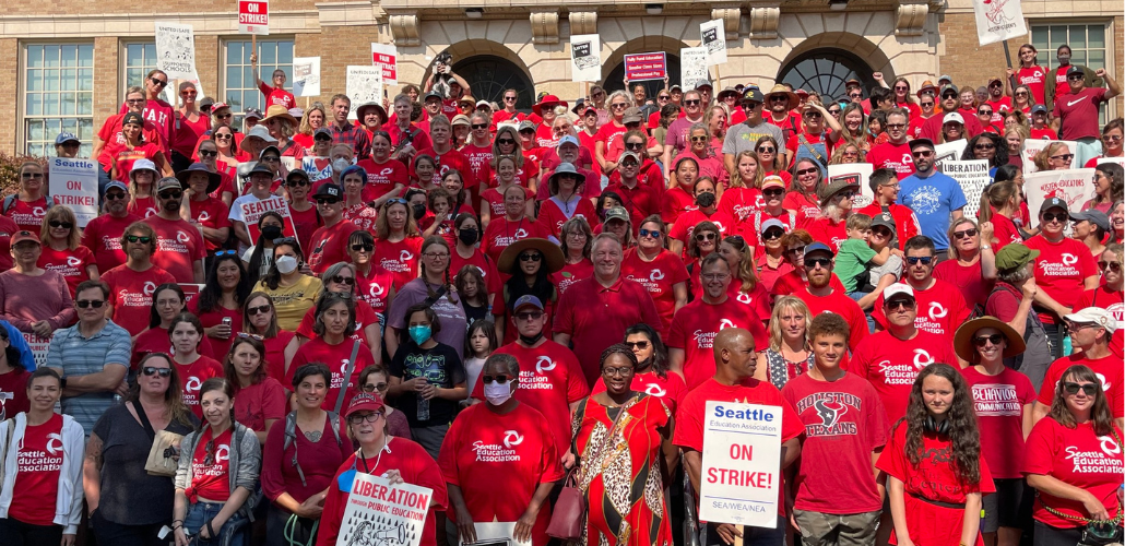 Large group of people outdoors, most wearing red shirts, some holding picket signs, on a sunny day. They are posed for a group photo on the steps of a school. The photo shows more than 200 people and it looks like there are more outside the frame.