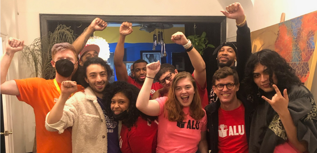 An exuberant group of young workers, diverse in race and gender, pose in a huddle and throw fists in the air