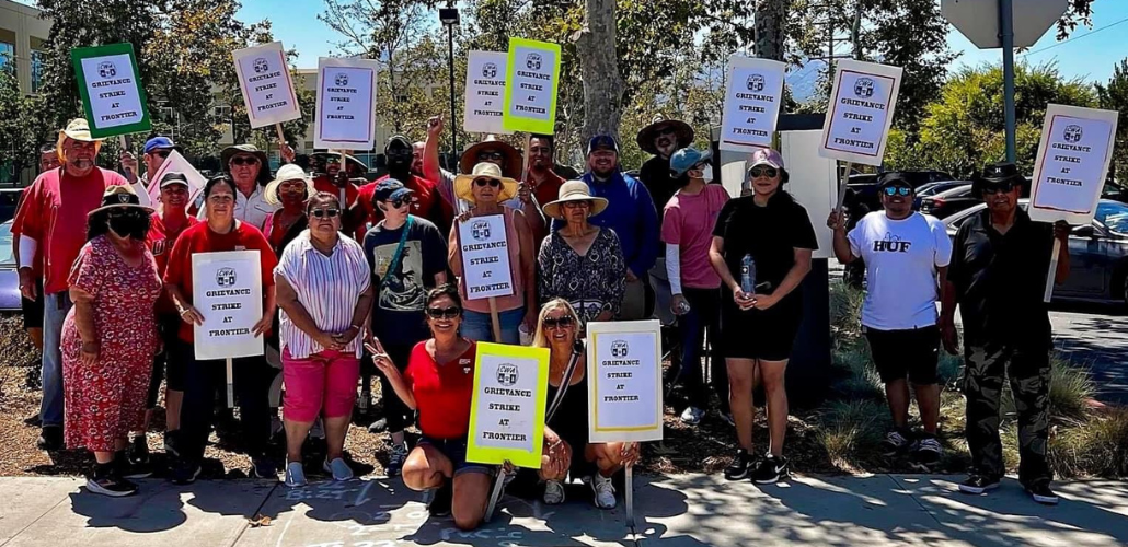 Crowd of people poses outside in the sun. Many carry signs with CWA logo that say "Grievance strike at Frontier."