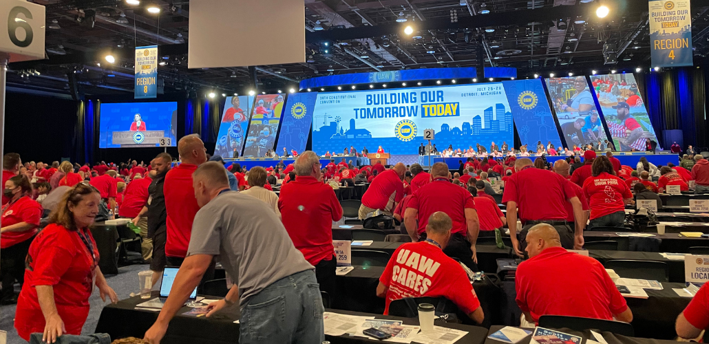 People in a big convention hall, all wearing red T-shirts, most getting to their feet. They are viewed from the back. At the front of hte room is a dais with people seated, and a huge lighted display that says "Building our tomorrow today" with the UAW logo.