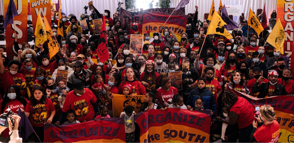 A diverse crowd of people, viewed from above, fills the frame. Most people are wearing red shirts. Many are Black. Many are lifting flags or raised fists and some hold "Organize the South" banners. Many little kids are in the front of the crowd.