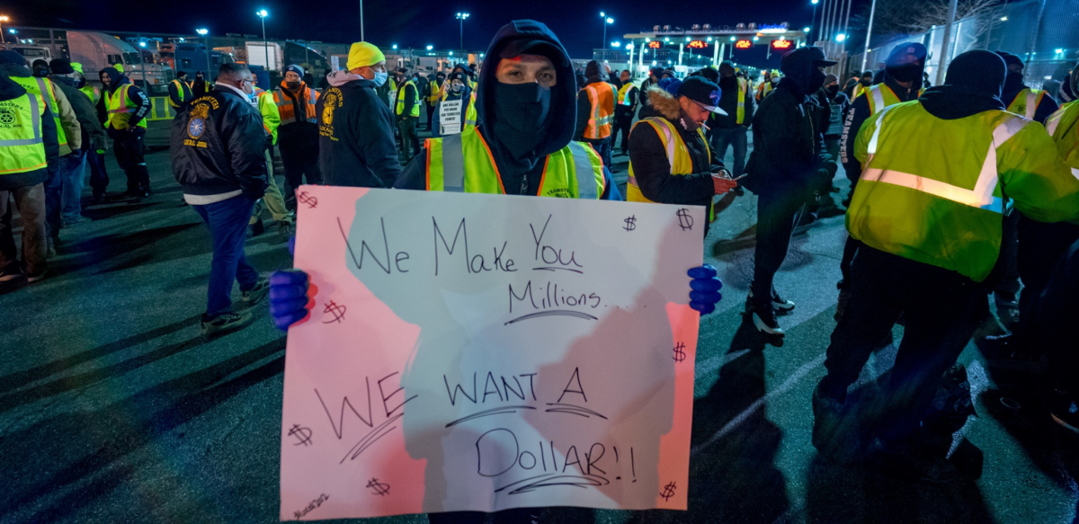 Person in yellow vest at night, outdoors in crowd, holds handwritten sign: "We make you millions, we want a dollar!"