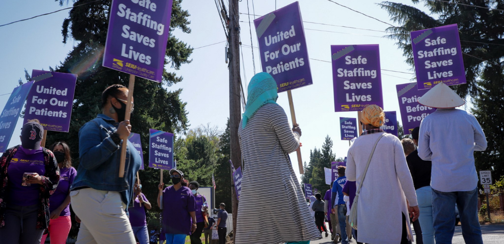 Workers picket in a circle. Many carry purple printed SEIUHealthcare signs that say "Safe Staffing Saves Lives" or "United for Our Patients." In the foreground is one worker viewed in profile and three silhouetted from behind; two of these workers wear head scarves and one wears the conical style of hat common in Southeast Asia.