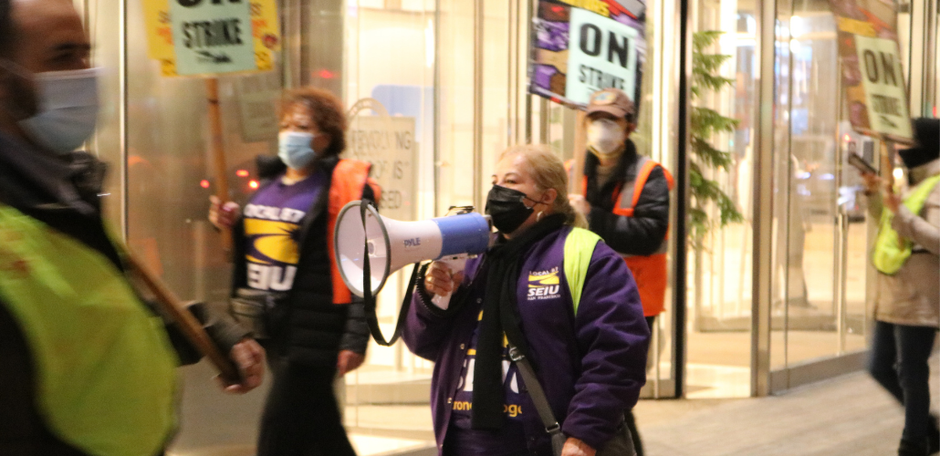 People in masks and purple SEIU 87 shirts picket in front of a bright window. Central person has a bullhorn. Others carry "ON STRIKE" signs and wear reflective vests.