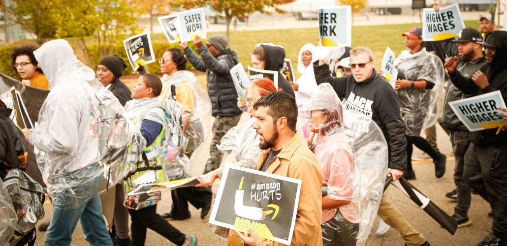 Workers, many in ponchos, march briskly on a street. Some carry printed signs saying "Safer work," "Higher wages," or "Amazon hurts."