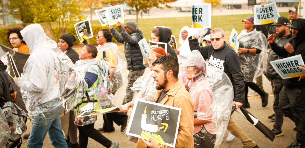 Workers, many in ponchos, march briskly on a street. Some carry printed signs saying "Safer work," "Higher wages," or "Amazon hurts." 