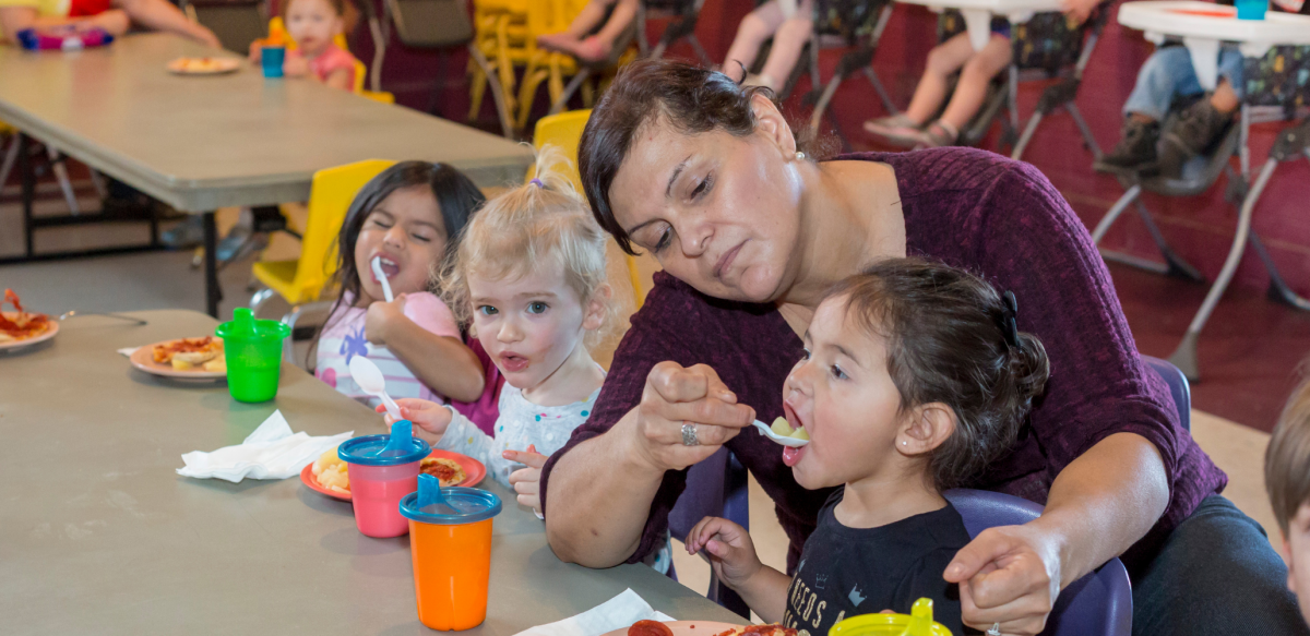 A woman spoon-feeds a toddler seated at a table next to two more toddlers, also eating. Visible behind her is another row of kids in high chairs.
