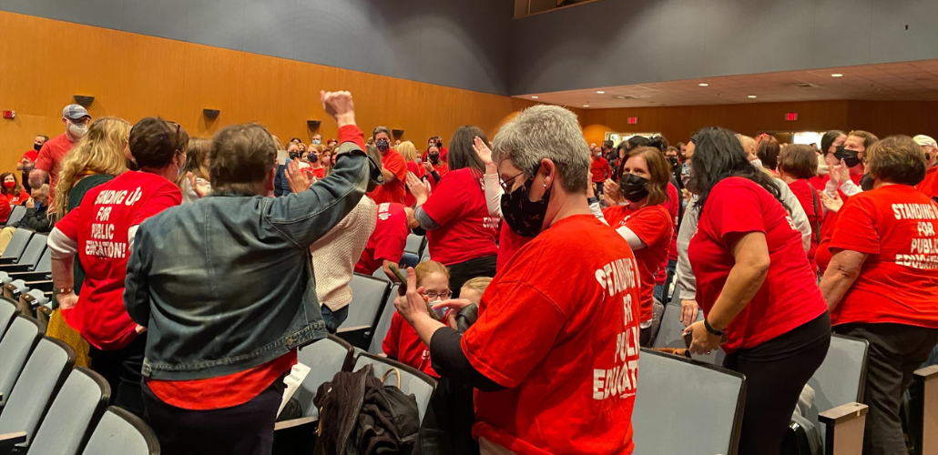 People, mainly women, in red T-shirts in the process of getting up en masse to exit an auditorium. One has a fist in the air, and several are clapping. Most are viewed from the back or side. The backs of their T-shirts say "STANDING UP FOR PUBLIC EDUCATION"