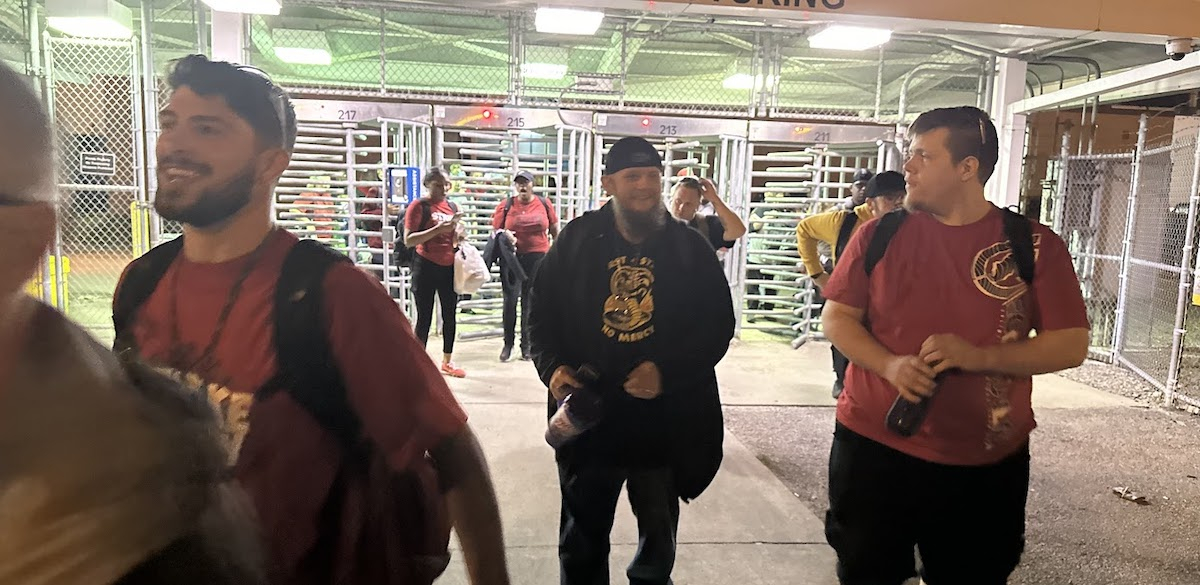 A group of smiling workers leave through factory turnstiles, walking towards the camera.