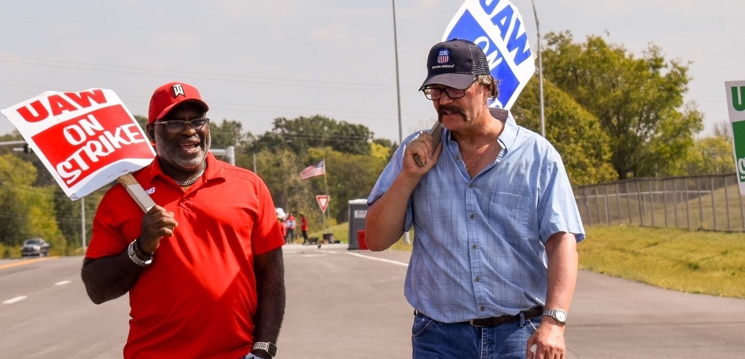 Two men, one Black and wearing red and one white wearing blue, walk together talking, each with a matching red or blue picket sign that says “UAW on Strike.”