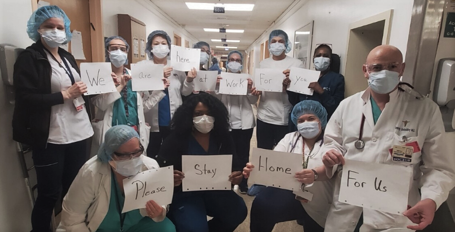 Masked health care workers in a hospital hold signs reading: "We... are... here... at... work... for.. you... Please... stay... home... for us."