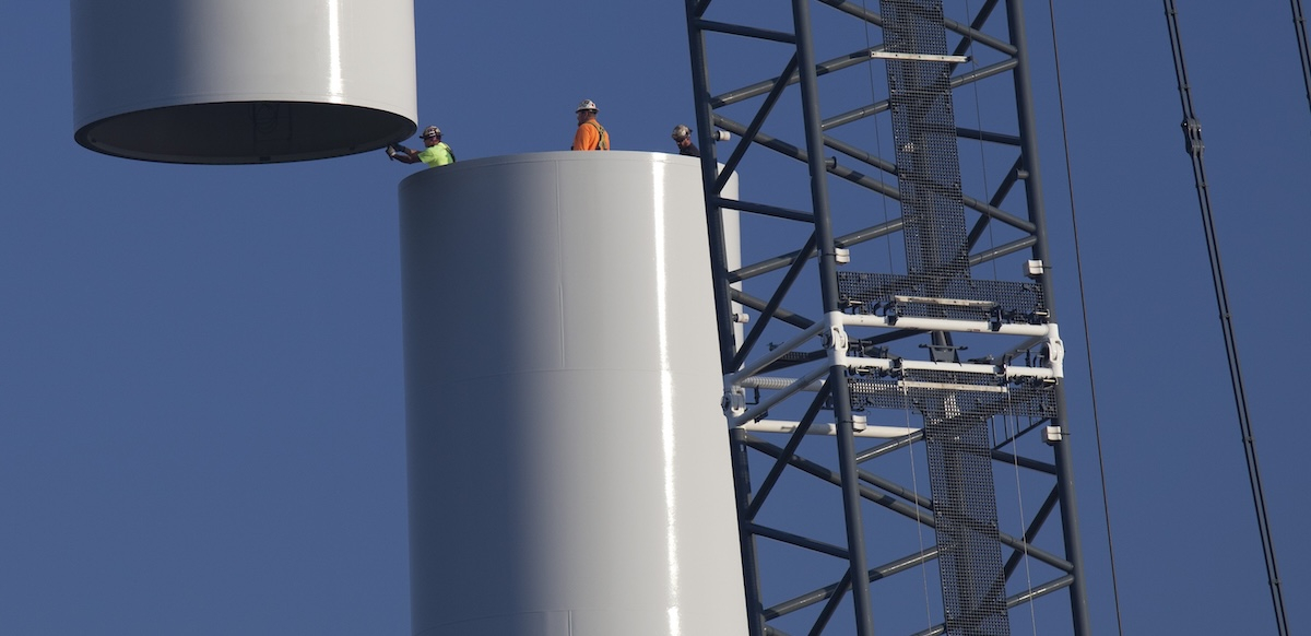 Workers join the top piece of a giant wind turbine to the bottom.  They are tiny against its hugeness.