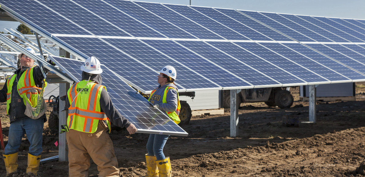 Three solar workers in yellow vests lift a large solar panel into place on a solar array.