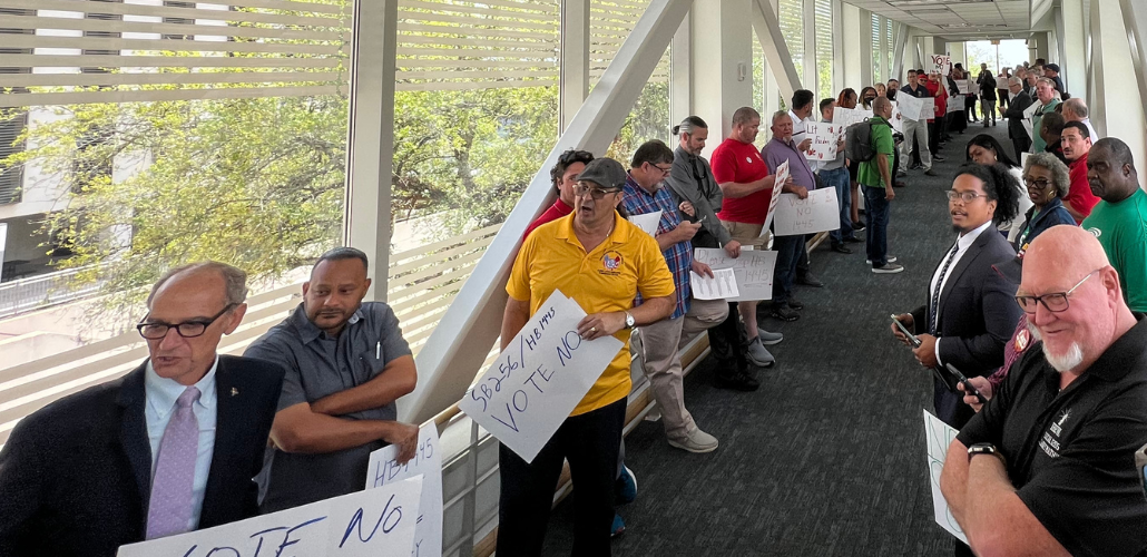 People in different colored union shirts or suits line both sides of a glass hallway. Many are men, some are Black and some are white. A few carry handmade signs urging a no vote on HB 1445.