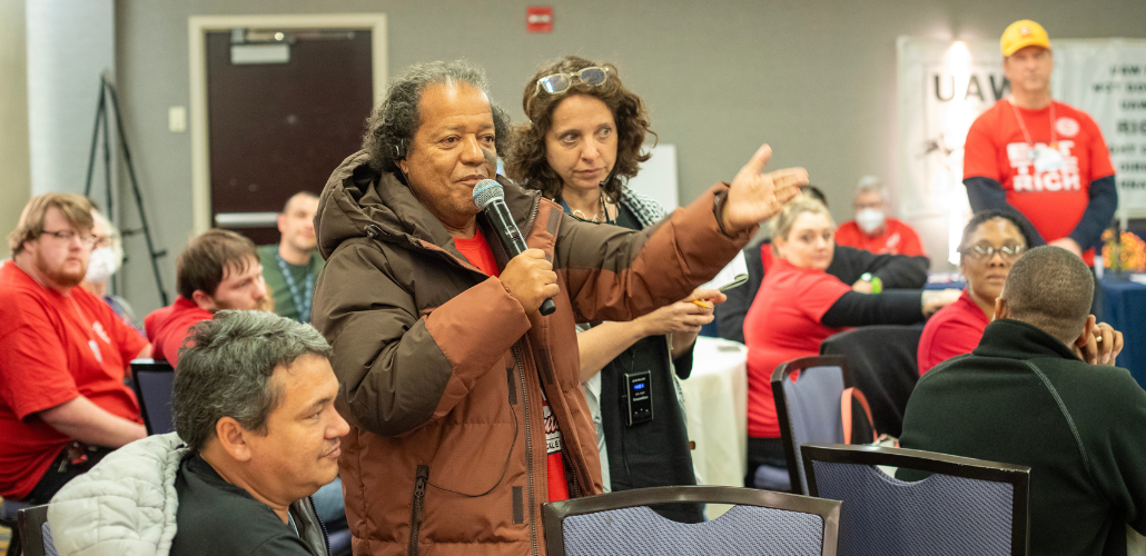 A person holding a mic stands speaking, gesturing with one arm, while others in the meeting listen intently. The group is diverse in race, gender, and age.