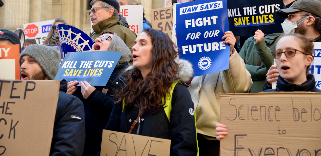People, mostly white, rally outdoors on building steps. Printed signs say "Kill the cuts, save lives" and "UAW says: Fight for our future!" Handmade signs can't all be read but one says "Science belongs to everyone."