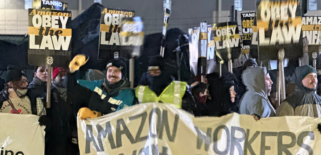 A crowd of workers stands outdoors in dim light. A smiling person with raised fist is one of several holding a handmade banner--the part visible says "Amazon Workers." Many people in crowd hold printed Teamster-logo picket signs that say: "Amazon: Obey the Law." Everyone is wearing coats with big hoods or warm hats; it looks cold out. Someone in lower left has a Staten Island T-shirt and keffiyeh