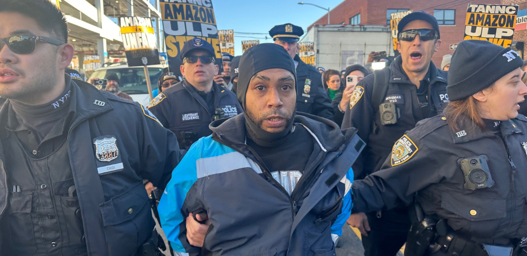 A man in a blue Amazon delivery driver jacket is flanked by bevy of cops, who are holding his arms and leading him away. More cops are behind him, and a crowd behind them. Printed Teamsters picket signs are visible saying "Amazon ULP strike" and "Amazon is unfair."