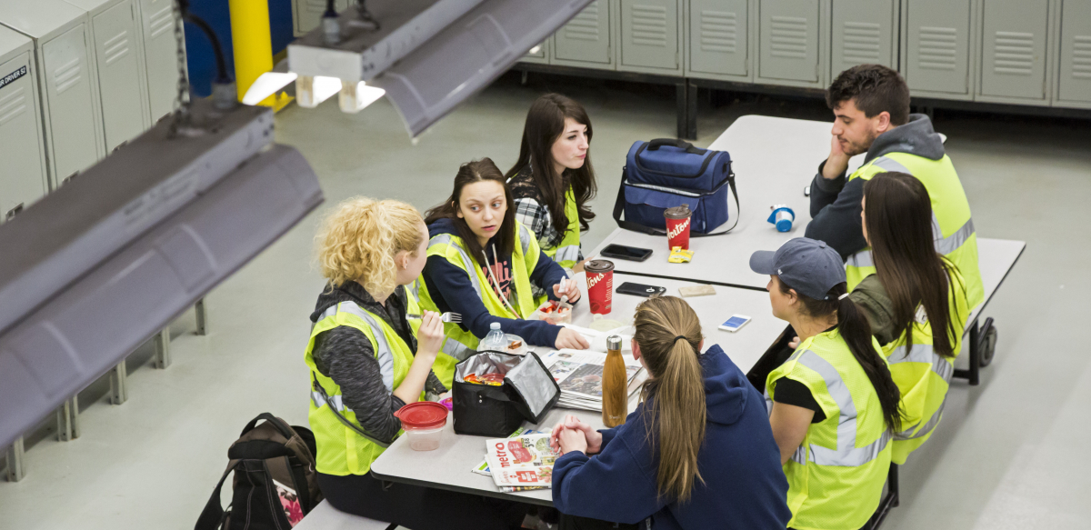 Workers in yellow vests sit around a lunch table at work.