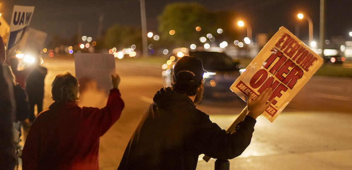 Workers hold signs at night--they are backlit by traffic and street lights but one sign is legible, saying ‘everyone tier one.’
