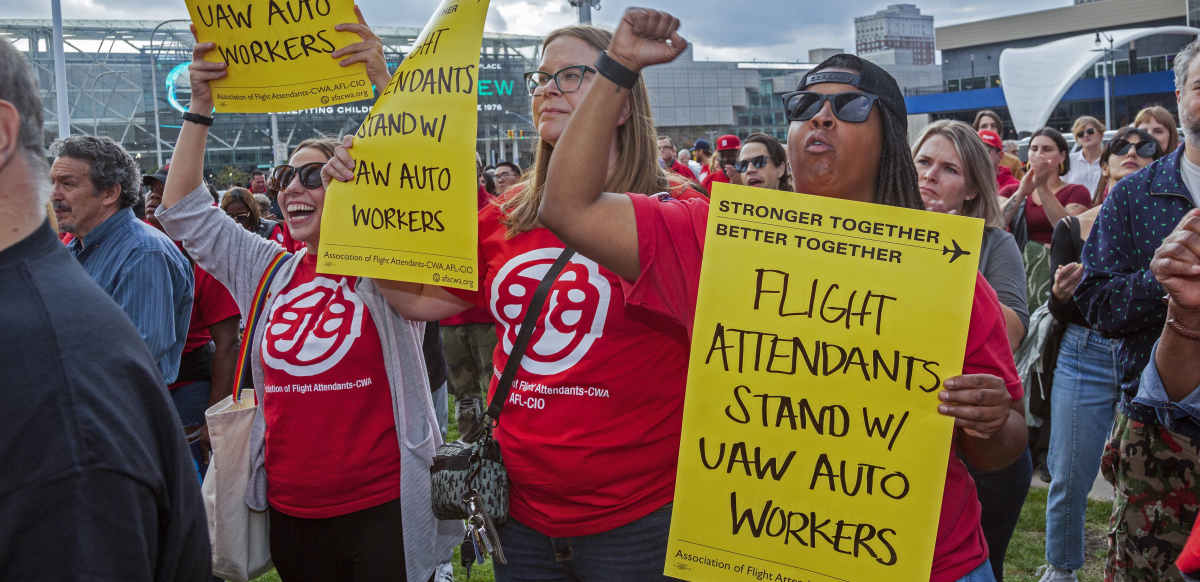 Three women wearing red Association of Flight Attendants shirts hold yellow signs saying, "Flight Attendants Support UAW Auto Workers."