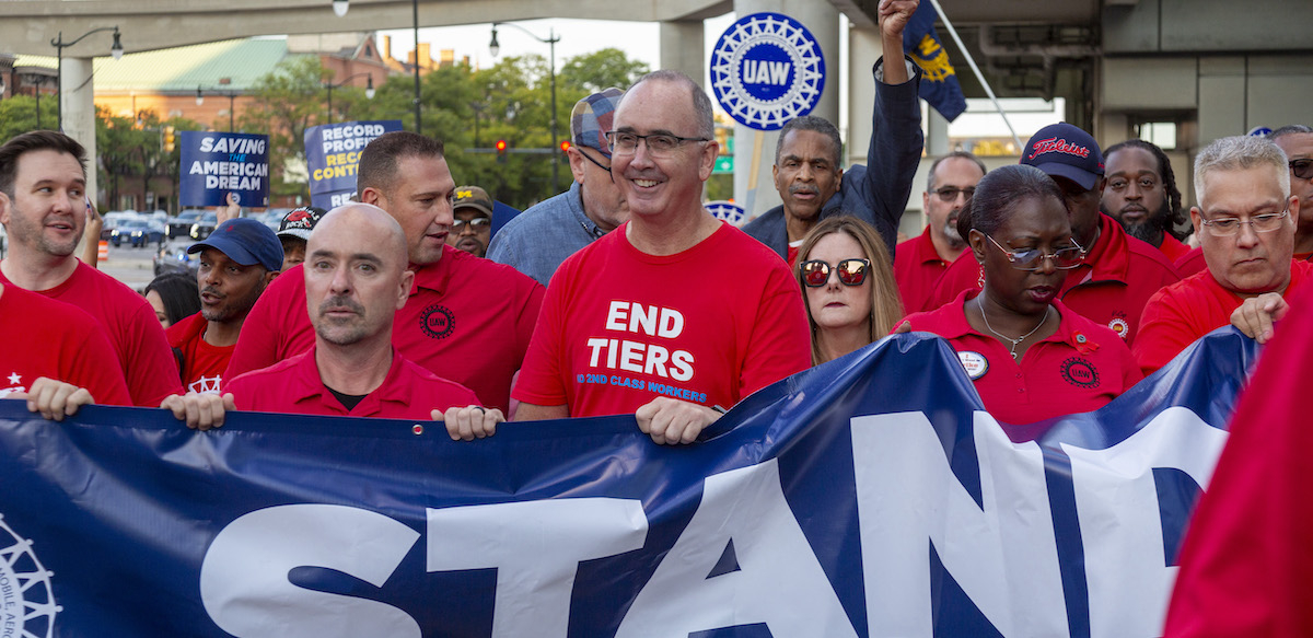 A group of men and women in red T-shirts stand holding a blue banner, the word ‘Stand’ is visible.