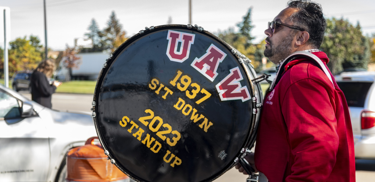 A UAW member walks with a drum that reads "UAW: 1937 - Sit-Down, 2023 - Stand Up."