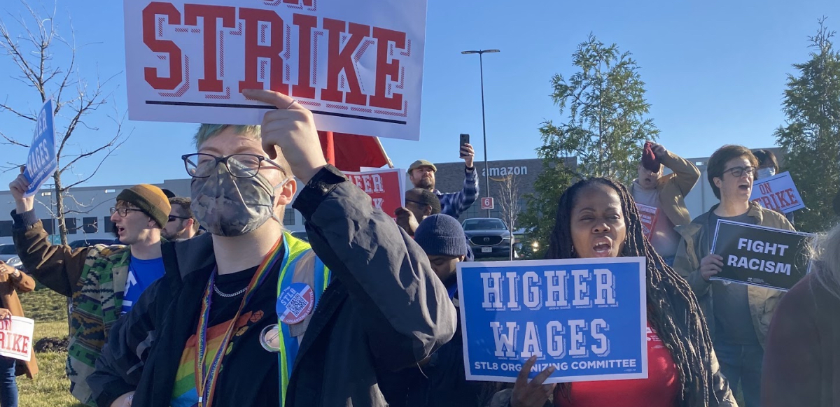 Amazon strikes hold signs saying strike, higher wages, stop racism. An Amazon facility is in the background