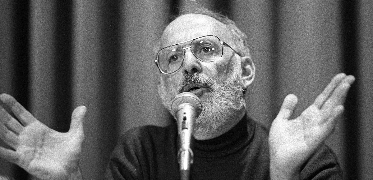 A bearded man speaks at a microphone with his hands wide open