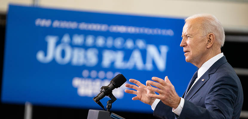 sideview of President Joe Biden speaking at a podium at Pittsburgh Carpenters Hall on March 31, 2021, with American Jobs Plan sign in background (white on blue)