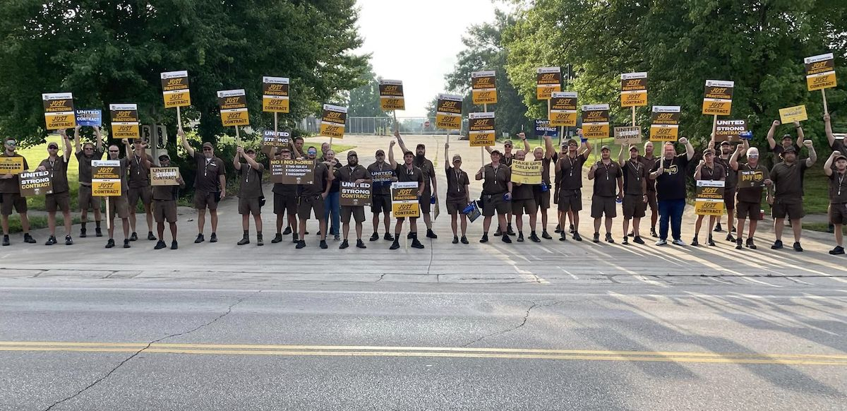 A row of people in UPS outfits with signs seen from far away block the road to some kind of facility, a gate is visible behind them.