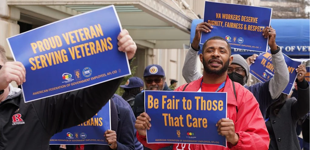 People (mainly Black men are visible) outside hold printed AFGE signs reading "Proud veterans serving veterans," "Be fair to those that care," and "VA workers deserve dignity, fairness, and respect."