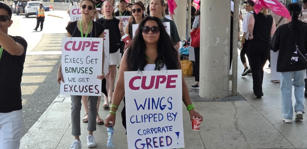Flight attendants, mainly women, march on an airport sidewalk. Some wave pink CUPE flags and others wear sandwich-board signs with "CUPE" printed on and handwritten slogans. The two most visible say "Wings clipped by corporate greed!" and "Execs get bonuses, we get excuses." The woman in the foreground meets the camera's gaze with a serious expression; women behind her are chanting or singing.