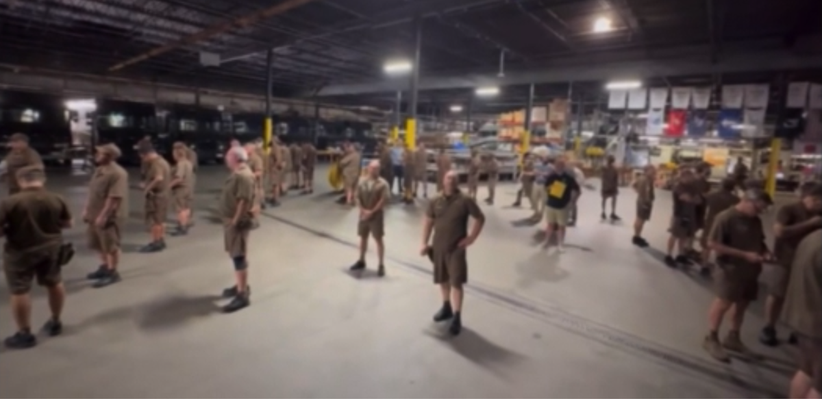 Indoors in a UPS warehouse, about 25 drivers are standing around the floor all with their backs to a manager with a yellow clipboard